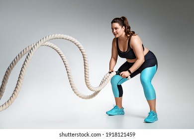 Strong woman working out with battle ropes. Photo of model with curvy figure in fashionable sportswear on grey background. Sports motivation and healthy lifestyle