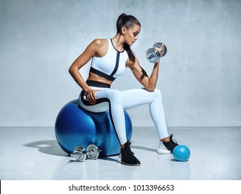 Strong woman working out with dumbbells sitting on gymnastic ball. Photo of sporty latin woman in sportswear on grey background. Sports