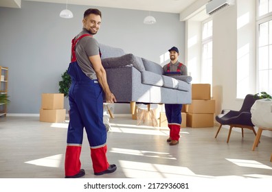 Strong Workers From Moving Company And Lorry Delivery Service Removing Furniture From Old Flat Or House. Two Happy Young Men In Workwear Uniforms Carrying Heavy Sofa Together