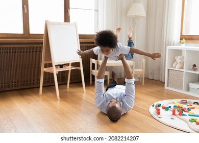 Strong Young African Dad Holding Little Preschool Son With Open Flying Hands, Lying On Heating Floor, Lifting Kid Up In Air. Father And Boy Playing Airplane Together, Doing Acroyoga, Enjoying Activity