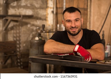 Strong Young Bearded Male Worker, Leaning With Both Hands On Newly Welded Metal Frame With Welder Machine. Handsome Man Wearing Black Shirt And Protective Gloves Smiling And Looking At Camera.