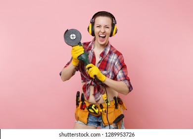 Strong Young Handyman Woman In Yellow Gloves, Noise Insulated Headphones, Kit Tools Belt Full Of Instruments Holding Power Saw Isolated On Pink Background. Female In Male Work. Renovation Concept