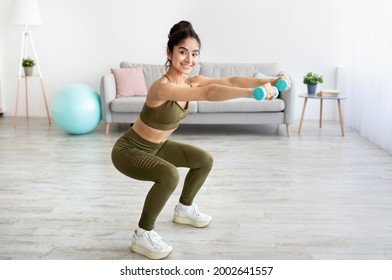 Strong young Indian woman working out with dumbbells, squatting at home, empty space. Fit Eastern lady taking care of her health, having domestic training, leading active lifesyle