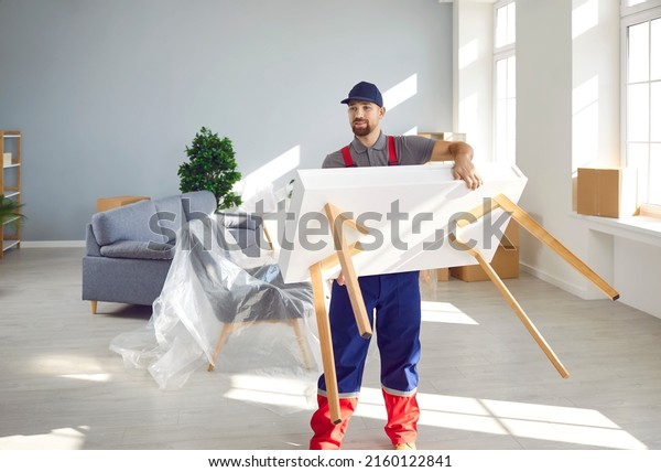 Strong young man from a moving company or\
truck delivery service carrying a white wooden table while removing\
furniture from a modern house or\
apartment