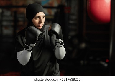 Strong young Muslim woman in boxing gloves, hijab and activewear looking at punching bag in gym and concentrating before hitting it - Powered by Shutterstock