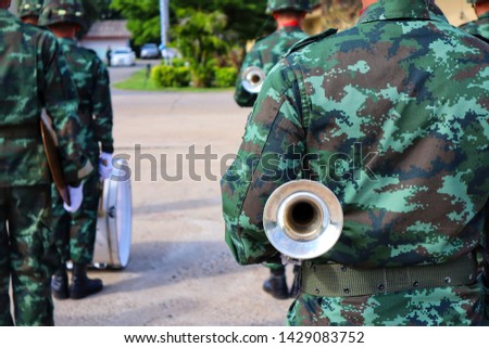 A strong young soldier stands upright, turning his back in an outdoor rowing practice. Musicians wait to perform.  Holding a trumpet instrument