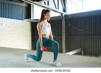 Strong young woman doing leg lunges while holding two dumbbells indoors. Fit and healthy woman wearing sportswear working out at the gym