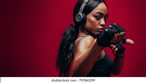 Strong young woman with headphones practising boxing. African female boxer exercising on red background - Powered by Shutterstock