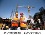 Structural engineer and architect dressed in orange work vests and  hard bats  discuss the construction process by the phone and use tablet  on the open building site near the crane