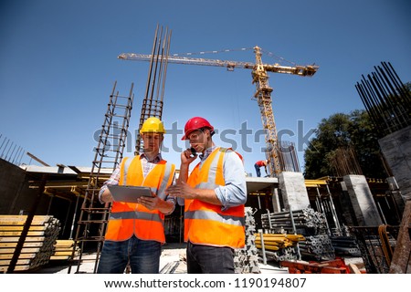 Structural engineer and architect dressed in orange work vests and  hard bats  discuss the construction process by the phone and use tablet  on the open building site near the crane
