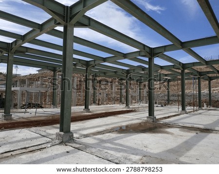 Structural steel frame of a building under construction against a bright blue sky. Low angle shot showcasing the green I-beams, concrete foundation, and scaffolding in the background. Perfect stock image for industrial development, civil engineering, commercial architecture, and construction project concepts.