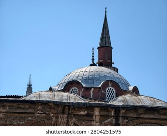 A structure features a rounded dome and a tall, pointed spire. Bright sunlight illuminates the building. Nearby tall buildings can be seen in the background.