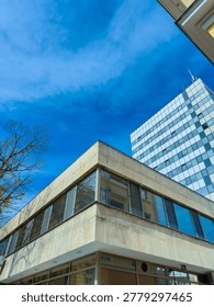 A structure has large windows that reflect the blue sky. Nearby buildings show a mix of modern design and older style. Trees are visible nearby, adding to the scene.