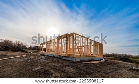 The structure of wood framing at construction site against a vast cloudy sky with bright sun behind. New build home at empty lot. framework ready for wall and roof install. Real estate development.