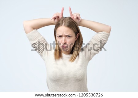 I am stubborn. Cute emotive woman with freckles pointing up with index fingers behind head as if it is horns, frowning over gray background.