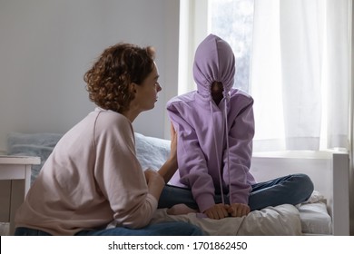 Stubborn teenage girl puts hood on head avoiding mom apology, ignoring talk with parent sitting on bed at home. Naughty teen daughter introvert hiding in hoodie showing disrespect to mother at home.