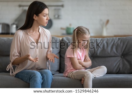 Stubborn upset little daughter ignoring strict young mother, sitting with arms crossed on couch in living room, angry mum lecturing unhappy preschool child family generations conflict concept