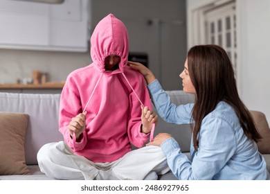 Stubborn upset teenage girl putting hood on head and avoiding her mom apology, ignoring talk with parent, sitting on couch at home - Powered by Shutterstock