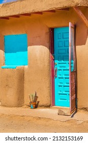 Stucco Taos Pueblo home with turquoise door and shutters and decorated flowerpot with feathers sitting outside - screen door held open with rock - all red clay