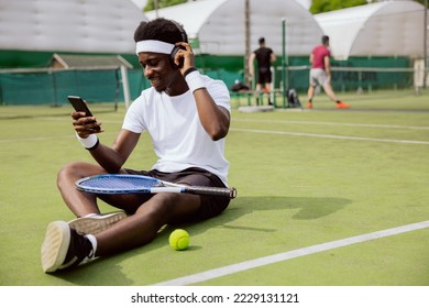 Student of African appearance is sitting on grass of tennis court, wearing headphones, holding phone and listening to music. Racket is on lap, and ball is on ground. He is dressed in sports clothes. - Powered by Shutterstock