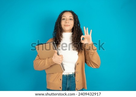 Student arab girl with curly hair smiling and looking happy, carefree and positive, gesturing victory or peace with one hand