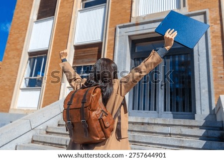 Student with backpack and folder raising arms in triumph, celebrating achievement and education milestones after finishing a course at a university campus
