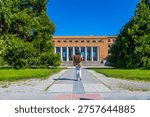 Student with a backpack walking on a pathway towards the main entrance of a university campus building on a sunny day, symbolizing education, learning, and future academic pursuits