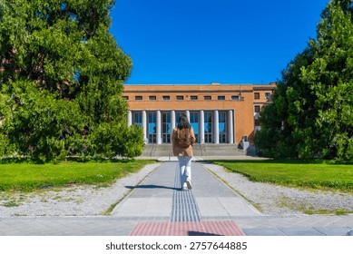 Student with a backpack walking on a pathway towards the main entrance of a university campus building on a sunny day, symbolizing education, learning, and future academic pursuits