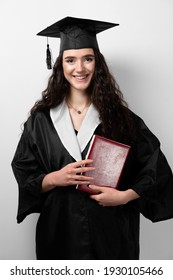 Student With Book In Graduation Robe And Cap Ready To Finish College. Future Leader Of Science. Academician Young Woman In Black Gown Smiling.