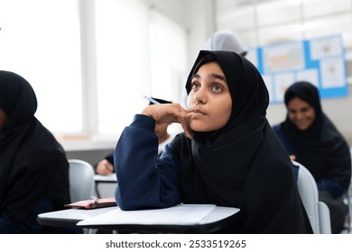 A student in a classroom, wearing a hijab, looks thoughtful. Classroom setting with students. Focused student, learning environment, education scene. Young Muslim girl wearing hijab in school. - Powered by Shutterstock