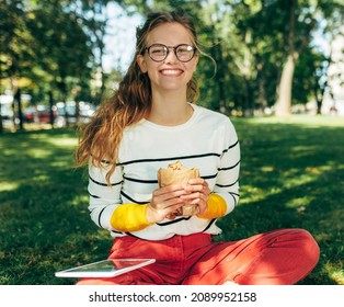 Student Female In Casual Outfit Waring Transparent Eyeglasses, Sitting On The Green Grass At The College Campus, Have A Lunch, And Studying. Woman Takes A Rest Eating Fast Food And Learning Outside.