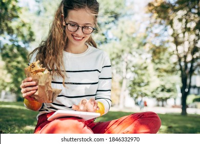 Student Female Sitting On The Green Grass The College Campus On A Sunny Day, Have Lunch And Studying. The Hungry Young Woman Takes A Rest Eating Fast Food And Learning In The Park.