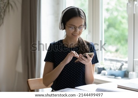 Student girl in headphones sit at table distracted from study holds smartphone hearing voice audio message from friend, enjoy free time use modern gadget, choose song, play music, tech, fun concept