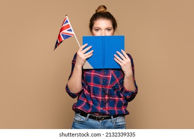 Student girl holds small UK flag on beige background. Young woman peeks out from behind open book, half face visible. Study abroad. International student exchange program. Learning British English. - Shutterstock ID 2132339335