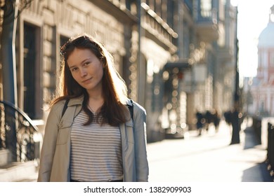 A student girl with long hair stands on the background of a city street with pedestrians, which is illuminated by the light of the sunset.