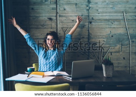 Student girl sitting at table with open notebook and raises hands up while smiling with sincere happiness. 
Woman overjoyed about successfully completed project on computer. Concept of achievement