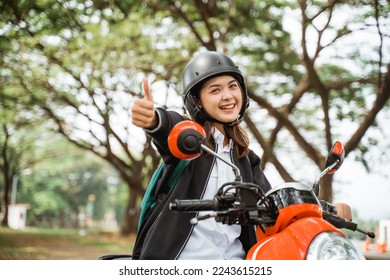 Student girl with thumbs up riding motorbike wearing helmet and jacket while going to school
