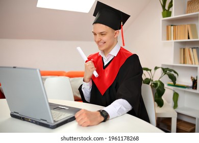 A Student In Graduation Gown And A Corner Cap Sits At Home In Front Of A Laptop During The Broadcast Of The Graduation Ceremony . Class Of 2021. Covid-19 And New Normal Concept.