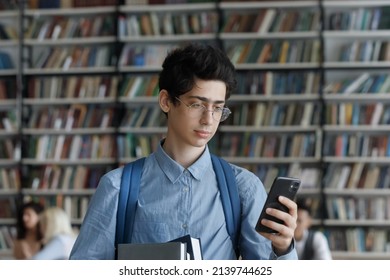 Student Guy In Glasses Holds Workbooks And Cellphone Check Messages, Chatting Online Standing In University Library. Gen Z Using Modern Tech And New Mobile Application, Class Timetable Apps Concept