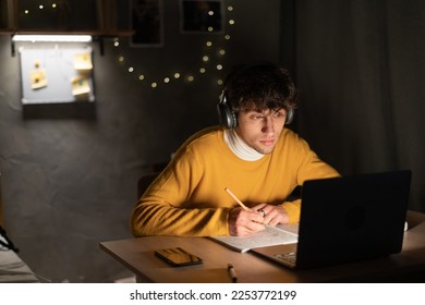 Student guy making notes in notebook using laptop computer sitting at desk at dormitory or home at night. Copy space