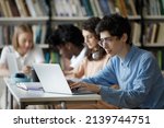 Student guy wear glasses studying in library or classroom, using laptop working on essay, prepare for college exams seated at table with diverse group mates. Education, professionals skills concept