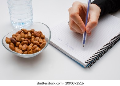 Student Hand Holding Pen And Writing Notes In The Notebook And Glass Bowl Of Hazelnuts And Water Bottle Isolated On White. Healthy Lifestyle