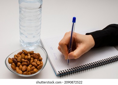 Student Hand Holding Pen And Writing Notes In The Notebook And Glass Bowl Of Hazelnuts And Water Bottle Isolated On White. Healthy Lifestyle