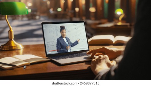 Student Learning From Online Lecture on Laptop in Library. African American Female Professor Teaching Engineering, Displayed on Screen, With Books and Notebook on Wooden Table