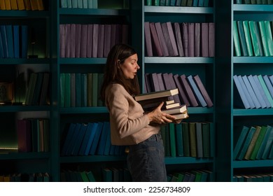 A student in the library, preparing for exams, studying at the university. A young woman holds a stack of heavy textbooks in her hands