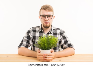 Student, Office And People Concept - A Young Grumpy Man Sitting At The Table With A Green Plant On White Background. He Looks Serious And Strange