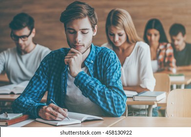 Student On Exam. Group Of Concentrated Young Students Writing Something In Their Note Pads While Sitting At Their Desks In The Classroom