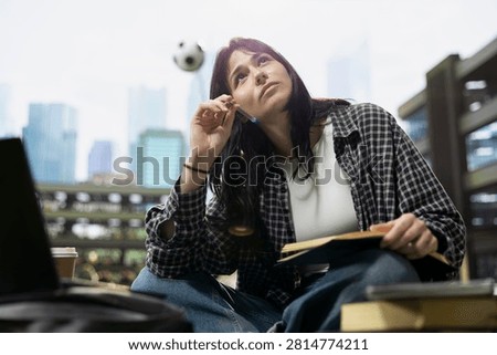 Student pauses while taking notes beside an open book and laptop on campus, looking up to consider ideas during independent study near stacked books in an outdoor academic setting.
