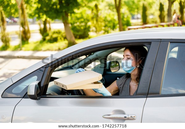 Student with pizza box in car with medical mask and\
gloves. Delivery food service drive by car. Safe food delivered\
according social distance. Tasty pizza with salami, cheese,\
tomatoes in cardoard\
box