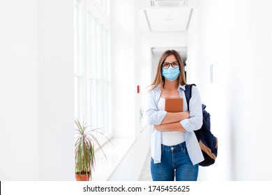 Student In Protective Face Mask In Empty College Indoors. Young Woman Going To Exams In High School. Girl With Backpack And Book In University Corridor During Quarantine, Lockdown.